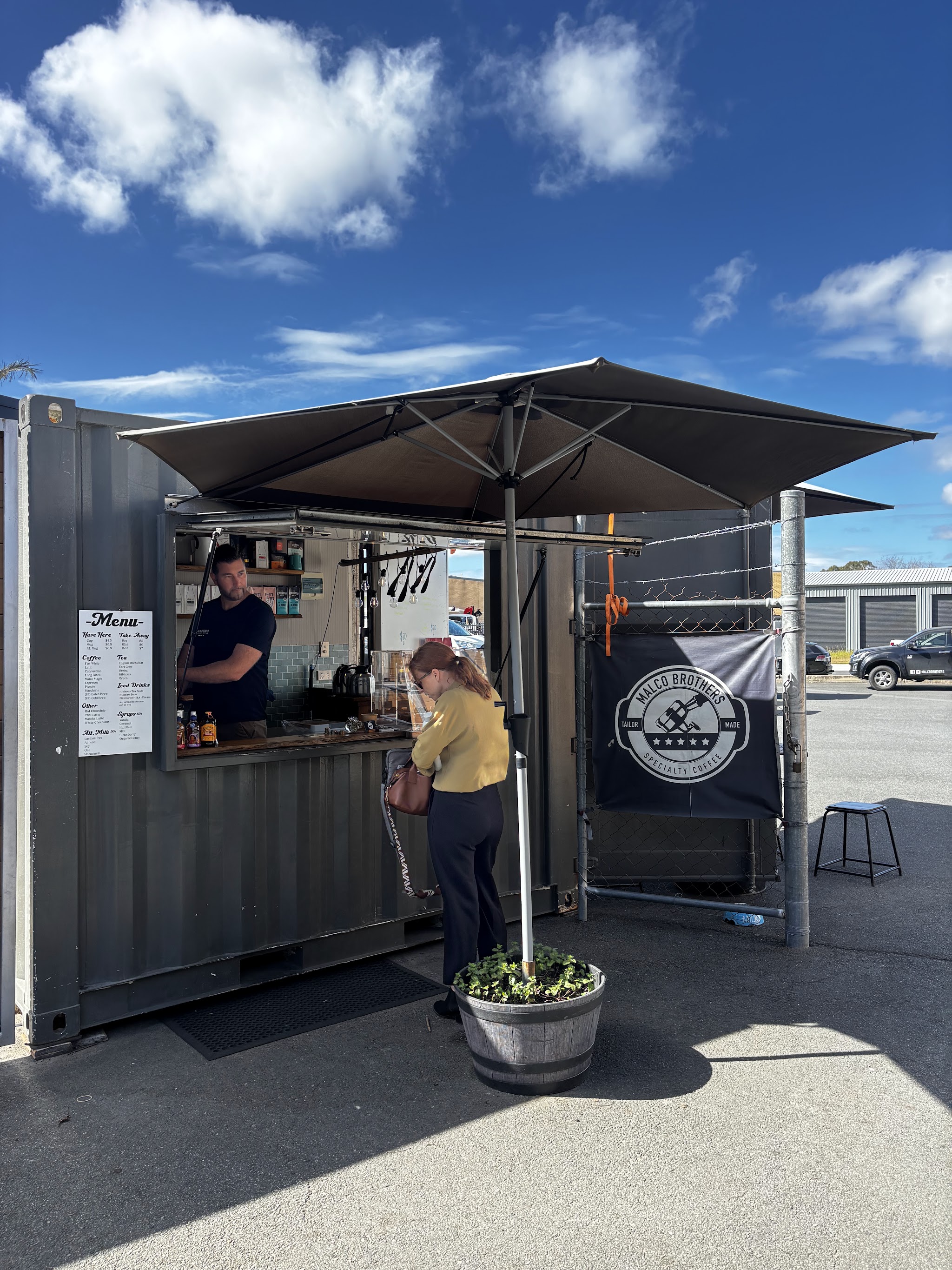 The barista serving a customer at the MALCO Brothers container window, with the menu board and roundel banner beside it